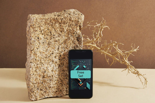 A black smartphone displaying a digital interface labeled 'Free Set' stands upright on a beige surface next to a large rough stone, with a dried branch against a brown backdrop.