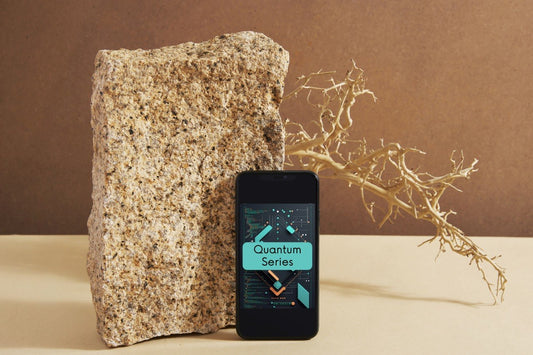 A black smartphone displaying a digital app titled 'Quantum Series' stands upright next to a large textured stone on a beige surface, with a brown backdrop and a dried branch behind it.